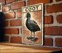 a wooden table with a framed illustration of a black bird, which appears to be a Coot, mounted on a brick wall.