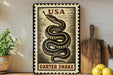 A framed image of a garter snake on a wall, with a wooden cutting board and a potted plant to the left.