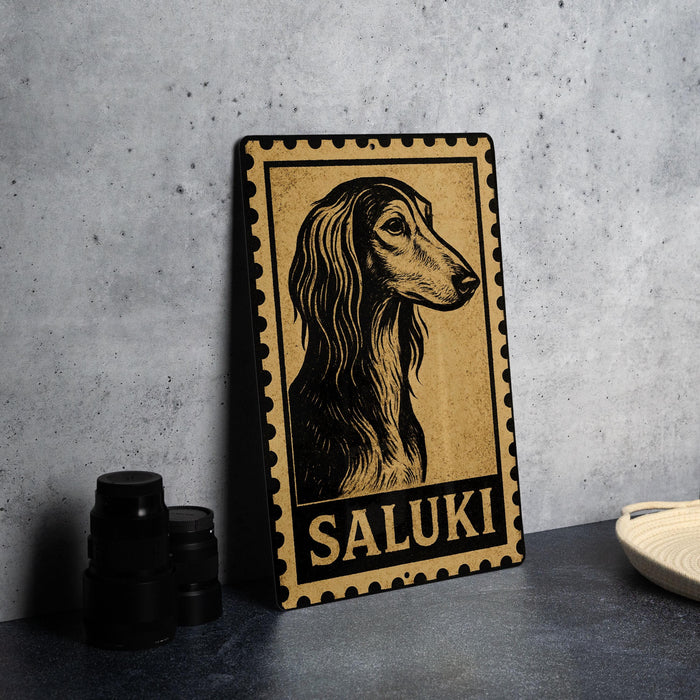 A metal sign with a stylized illustration of a dog's head and the word "Saluki" printed on it, placed on a gray countertop next to some black camera lenses.