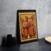 A framed illustration of two cartoon pigs is displayed on a dark surface, with a stack of black camera lenses and a white dish visible to the side.