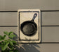 a sign with a black and white illustration of a frying pan on it, mounted on a wall.