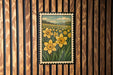 A framed illustration of a field of yellow daffodils is displayed on a wooden wall.