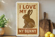 a sign with the text "I LOVE MY BUNNY" and an illustration of a rabbit. The sign is placed on a shelf next to a wooden cutting board and a potted plant.