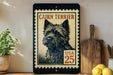 A framed image of a cairn terrier dog, with a postage stamp-like border, is displayed on a shelf next to a wooden cutting board and some lemons.