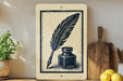 a wooden board with a black and white illustration of a quill pen and inkwell on it, placed on a wooden surface next to a potted plant and a wooden cutting board.