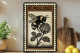 a framed illustration of a bumble bee on a flower, placed on a shelf next to a wooden cutting board and some lemons.