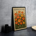 A framed illustration of a bed with orange and yellow flowers, resembling a postage stamp design, is displayed on a table.