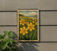 a framed painting of a field of yellow daffodils against a backdrop of green grass and hills.
