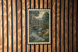 A framed illustration of a wooden bridge spanning a river is displayed on a wooden wall.