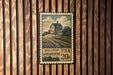 A vintage-style postage stamp featuring a farmhouse illustration, mounted on a wooden wall.