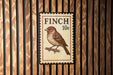 A postage stamp-like sign with a brown finch illustration and the text "FINCH 10c" is displayed on a wooden wall.