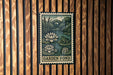 A metal sign with a stamp-like design hangs on a wooden wall, depicting a garden pond with a frog and water lilies.