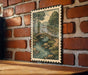 A framed illustration of a bridge over a river is hanging on a brick wall.