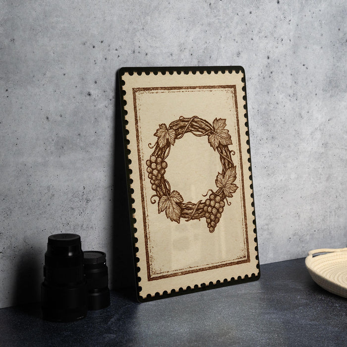 a framed picture of a wreath with grapes on a table, accompanied by a stack of black camera lenses and a white dish.