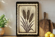 a decorative tile with a wheat design, placed on a shelf next to a wooden cutting board and a potted plant.
