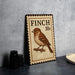 A metal sign with a bird illustration and the text "FINCH 10c" is displayed on a table.