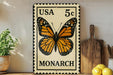 A framed US 5c postage stamp featuring a detailed illustration of a monarch butterfly is displayed on a wooden shelf, accompanied by a wooden cutting board, a potted plant, and some lemons.