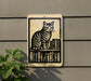A sign featuring a cat sitting on a bookshelf, with a plant to the left of the sign.