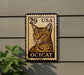 A wooden postcard-style stamp featuring a cat's face, mounted on a wall next to a plant.