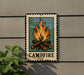 A metal sign with a campfire illustration on it, mounted on a wall.