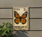 A US 5c postage stamp featuring a detailed image of a monarch butterfly is displayed on a wall.