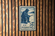 A blue and white postage stamp featuring a person fishing, with the text "ICE FISHING 26" printed on it.