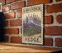 A vintage-style metal sign with a floral illustration of lavender flowers and the word "HEDGE" is displayed on a brick wall.