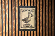 A sign featuring a goose on a postage stamp-like background, mounted on a wooden wall.
