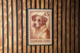 A brown and white dog is depicted on a brown and white postage stamp with the number 45, which is affixed to a wooden wall.