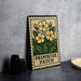 A framed print of a yellow flower with green leaves, labeled "Primrose Patch", is displayed on a table next to a stack of black camera lenses.