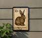 A 10 cent postage stamp featuring a rabbit illustration, mounted on a wall next to a plant.