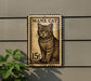 A vintage-style postage stamp featuring a cat illustration, mounted on a wall next to a plant.
