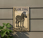 a zebra standing on a patch of grass, with the text "PLANS ZEBRA EQUUS QUAGGA" displayed on the sign.