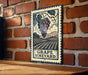 A vintage-style metal sign with a grape vineyard illustration is mounted on a brick wall.