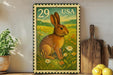 A framed stamp depicting a rabbit in a field of flowers, with the number 29 and the word "USA" printed on it. The stamp is placed on a shelf next to a wooden cutting board and a potted plant.
