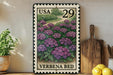 A framed postage stamp depicting a garden scene with purple flowers, placed on a wooden surface next to a wooden cutting board and some potted plants.