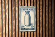 A vintage-style postage stamp featuring a milk bottle, mounted on a wooden wall.