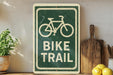 a green and white sign that reads "BIKE TRAIL" with a bicycle symbol, placed on a wooden surface next to a potted plant and a wooden cutting board.