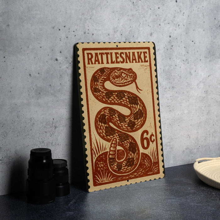 A metal sign with a printed design of a rattlesnake, resembling a postage stamp, is displayed on a table.