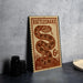 A metal sign with a printed design of a rattlesnake, resembling a postage stamp, is displayed on a table.