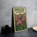 A metal sign with a floral design, featuring the text "PHLOX GARDEN" and a postage stamp-like border, is displayed on a table next to some black camera lenses.