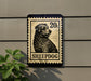 A metal sign with a black and white illustration of a dog, resembling a postage stamp, mounted on a wall.