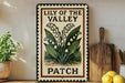 A framed sign with the text "Lily of the Valley PATCH" is displayed on a shelf next to a wooden cutting board and a potted plant.