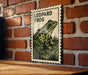 A framed illustration of a leopard frog is displayed on a brick wall.