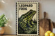 A framed illustration of a leopard frog is displayed on a shelf, accompanied by a wooden cutting board and a potted plant.