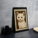 A framed image of a cat is displayed on a black surface, with a stack of black camera lenses and a white dish visible to the right.
