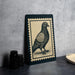 a framed illustration of a pigeon on a black background, placed on a table next to some black camera lenses.