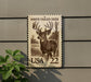A white-tailed deer stamp with the text "WHITE-TAILED DEER" and "USA 22" printed on it, mounted on a wall next to a plant.