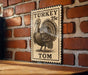 A wooden table with a brick wall in the background, featuring a vintage-style metal sign with a turkey illustration and the text "TURKEY 25c TOM".