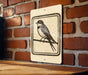 A sign featuring a black and white illustration of a bird perched on a branch is mounted on a brick wall.
